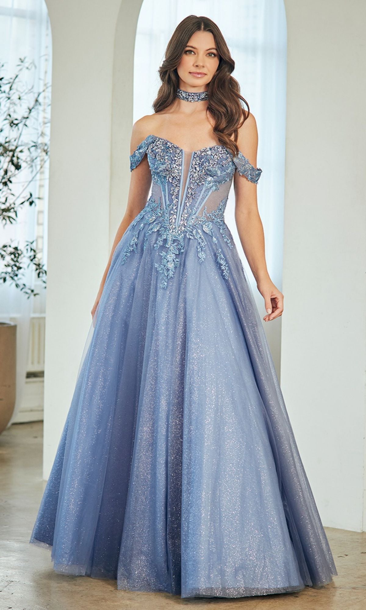 A woman stands indoors wearing the Amelia Couture 8065—a floor-length, off-the-shoulder prom dress with a corset bodice, shimmery blue glitter tulle, floral embellishments, and a matching choker.